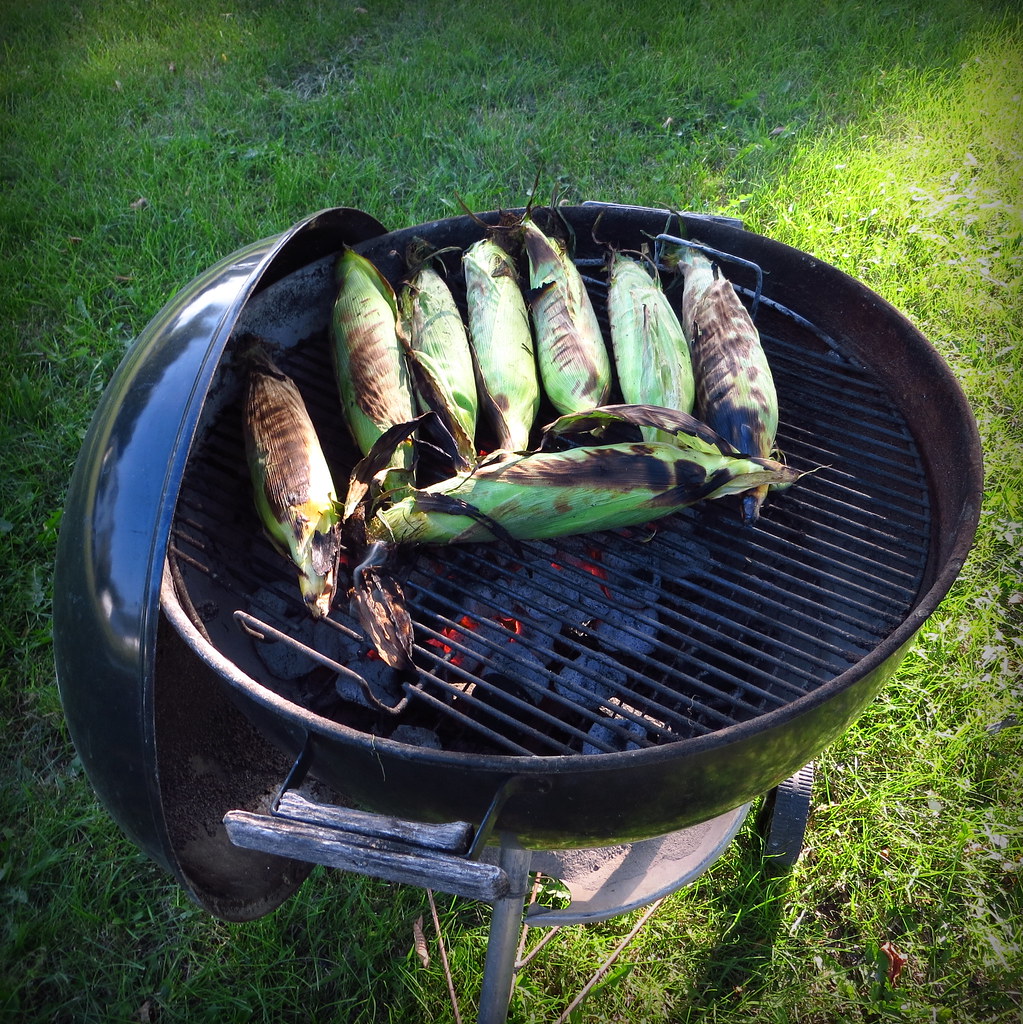Sweet corn cobs roasting in their husks across an open charcoal grill